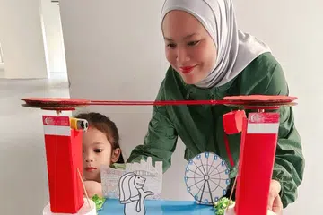 Cik Shairah Shazana Mohamed (kanan) bersama anak bongsunya berusia empat tahun, Clemira Wahyudi Ichwandy bergambar dengan hasil kraf Hari Kebangsaan mereka iaitu replika kereta kabel Singapura yang bergerak.