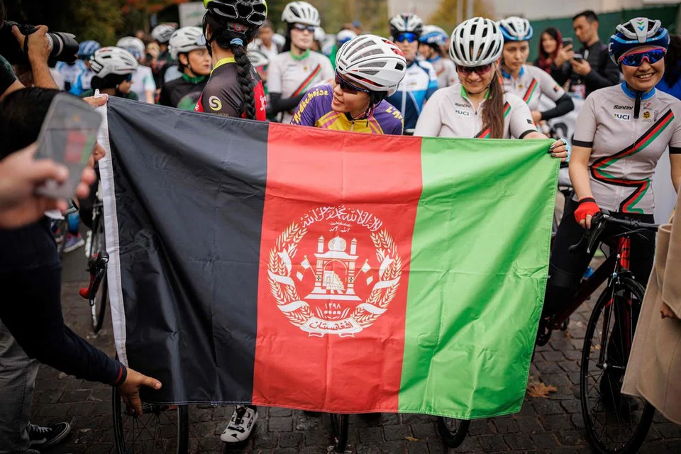 Masomah Ali Zada (tengah), yang merupakan peserta lumba basikal Afghanistan pertama yang menyertai sukan Olimpik sebagai seorang anggota Jawatankuasa Olimpik Antarabangsa pasukan pelarian, menanti di belakang bendera Afghanistan semasa permulaan kejohanan lumba basikal jalan raya 2022 di  Aigle, barat Switzerland pada 23 Oktober 2022. 