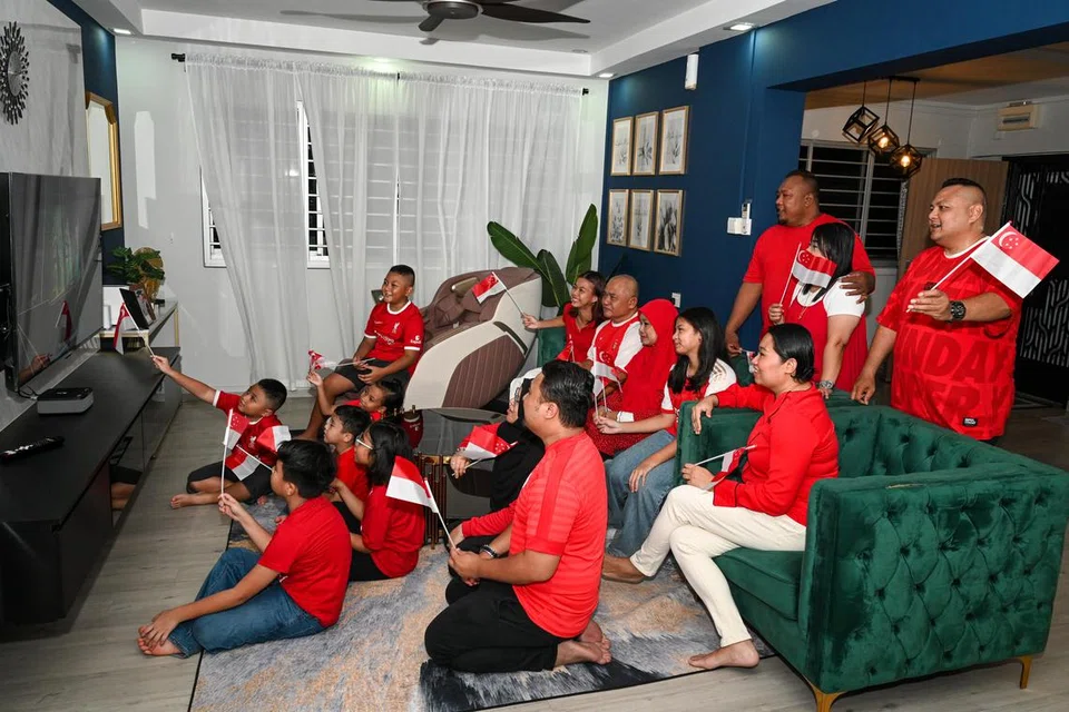 Encik Muhammad Shahid Haron (berseluar hitam duduk di lantai) bersama keluarga dan jiran tetangganya meraikan ulang tahun Singapura dengan menonton Sambutan Kejiranan Hari Kebangsaan secara langsung melalui platform penstriman di kaca televisyen. Sambutan itu diadakan di padang terbuka bersebelahan pusat beli-belah Waterway Point di Punggol pada 10 Ogos.