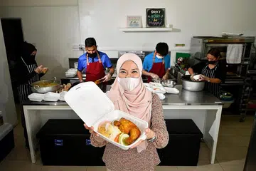 HIDANGAN SEGAR BAGI KELUARGA ISTIMEWA: Cik Faraliza Zainal menunjukkan sebungkus makanan yang akan diberikan kepada keluarga berpendapatan rendah yang mempunyai kanak-kanak berkeperluan khas. Makanan-makanan itu dibungkus oleh pekerja berkeperluan khas di Ashraf's Cafe (belakang). - Foto BH oleh LIM YAOHUI