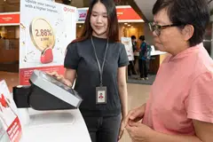 Sebagai seorang duta digital di OCBC, peranan Cik Siti Hatijah (kiri) termasuk membantu pelanggan di mesin ATM, membimbing mereka menggunakan perbankan digital, serta menyokong operasi cawangan. 
