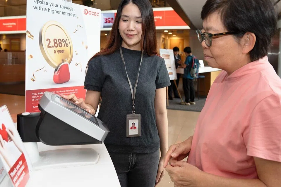 Sebagai seorang duta digital di OCBC, peranan Cik Siti Hatijah (kiri) termasuk membantu pelanggan di mesin ATM, membimbing mereka menggunakan perbankan digital, serta menyokong operasi cawangan. 