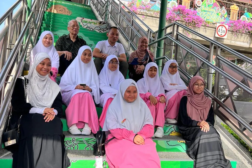 Mayor CDC South East, Encik Fahmi Aliman (barisan atas, tengah) bersama Pengerusi Lembaga Pengurusan Cik Ma’mun Suhaimi (barisan atas, kiri), Pengetua Madrasah Al-Ma’arif Al-Islamiah, Cik Maisarah Kasim (atas, kanan) bersama pelajar yang terlibat berfoto di tangga jejatas di mana karya mereka dipamerkan. 