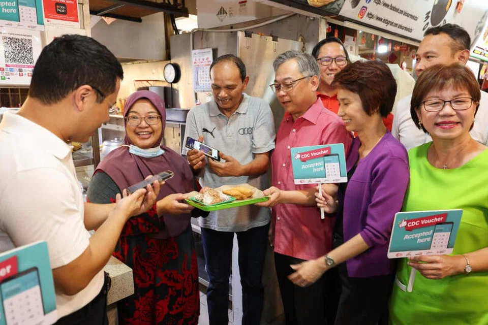 Menteri Pembangunan Negara, Encik Desmond Lee (kiri) menggunakan baucar CDC untuk membeli epok-epok dan ondeh ondeh daripada pemilik gerai 722 Malay Nasi Padang di West Coast pada 25 Jun. Beliau diiringi Timbalan Perdana Menteri, Encik Gan Kim Yong (barisan depan, tiga dari kanan); Mayor CDC North West, Encik Alex Yam (belakang, dua dari kanan); Menteri Negara Kanan (Perdagangan dan Perusahaan), Cik Low Yen Ling; Mayor CDC South East, Encik Mohd Fahmi Aliman (belakang, paling kanan) dan Mayor Daerah Central Singapura, Cik Denise Phua (paling kanan).