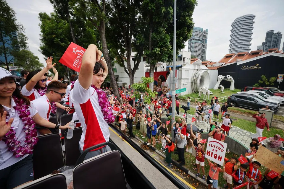 Dalam memaparkan gelagat lucunya, atlet luncur lelayang, Maximilian Maeder (kanan) membuat isyarat berbentuk hati kepada para penyokong semasa perarakan sambutan One Team Singapore melalui Victoria Street pada 14 Ogos. Turut bersama dalam sambutan di Victoria Street adalah kedua ibu bapa Maeder, iaitu Encik Valentin Maeder dan Cik Teng Hwee Keng (kededuanya terselindung).