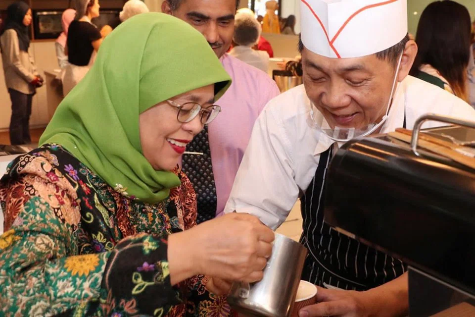 GEMBIRA BERINTERAKSI: Presiden Halimah Yacob sempat mempelajari cara membuat kopi daripada Encik Chua Kok Leong, seorang sukarelawan yang mengendalikan kafe 'Give n Take' di Pusat Aktiviti Sasco@Khatib. - Foto MCI