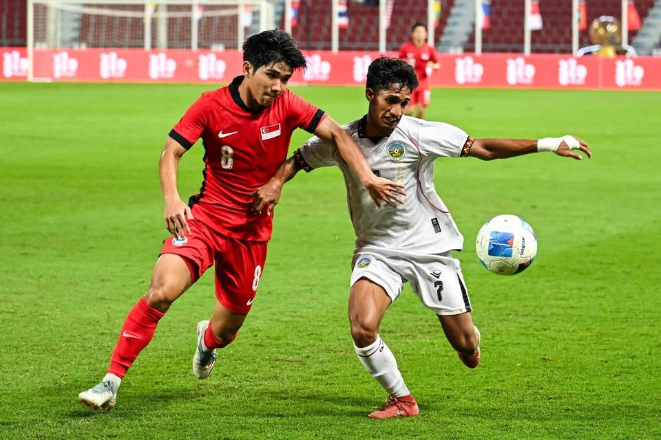 Pemain Singapura, Andrew Aw (jersi merah), kelihatan sedang berebut bola dengan pemain Timor-Leste, Luis Figo. 