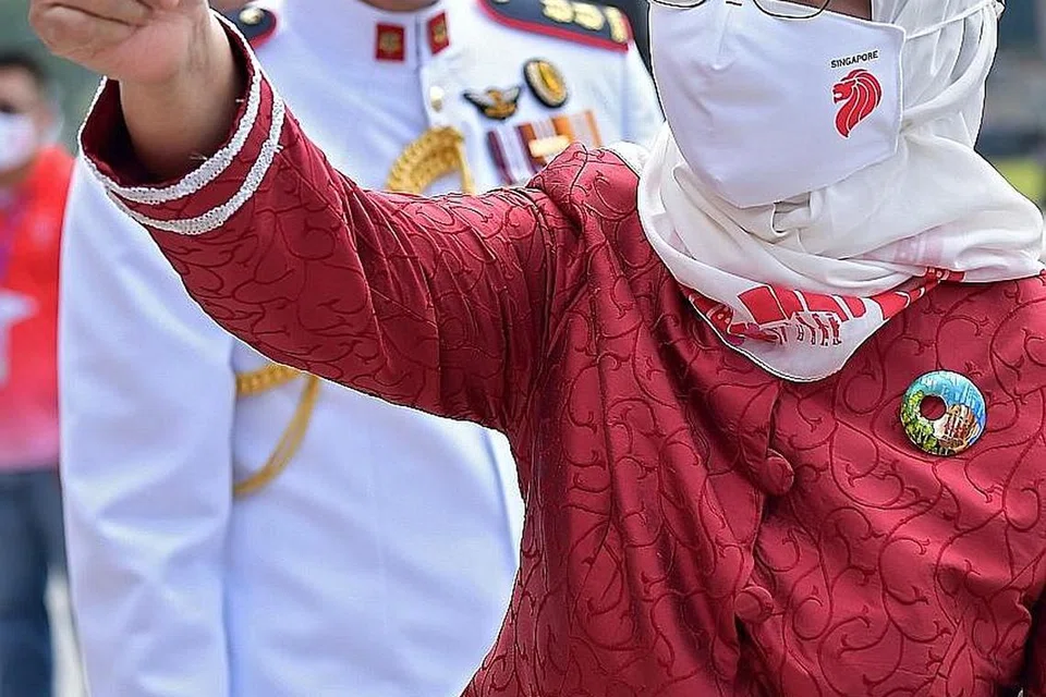 MERAH PUTIH: Presiden Halimah Yacob (gambar kanan), yang menggayakan kerongsang enamel dan selendang putih yang rekaannya mendapat inspirasi daripada hasil seni artis kurang upaya dan dicipta bersama jenama setempat di bawah inisiatif SG Enable, memberi isyarat jempol kepada penonton setibanya di Padang semalam. - Foto-foto BH oleh NG SOR LUAN.