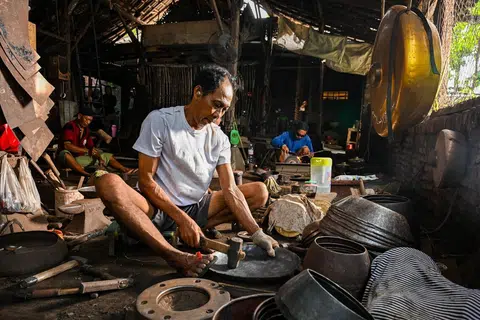 Tiga petukang sedang bertugas di stesen masing-masing dengan setiap seorang menumpukan perhatian pada peringkat berbeza dalam proses menghasilkan gamelan.