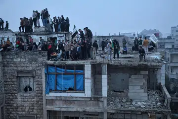 Orang ramai berdiri di atas bumbung bangunan yang sebahagiannya rosak ketika perarakan sempena ulang tahun pertama penyingkiran mantan Presiden Syria Bashar al-Assad, di Idlib, Syria, pada 8 Disember 2025. 