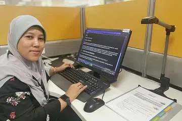 TIADA HALANGAN LAKUKAN KERJA: Cik Nuraziana Mohamed Said melakukan tugasnya dengan bantuan alat seperti pengimbas khas dan sofwe yang membacakan perkataan. - Foto MCCY