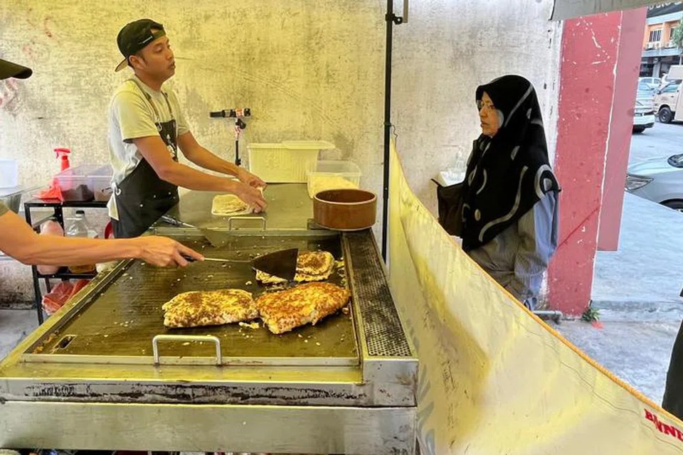 Peniaga, Encik Mohd Daniel Shukor, 34 tahun (kiri depan), melayani pelanggannya, Cik Zaimah Omar, 64 tahun (kanan depan), di gerainya  ‘Murtabak Singapore Dari Rumah’.