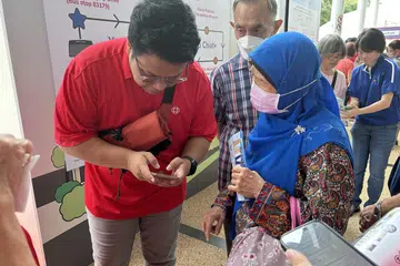 Cik Rahmah Abdullah (kanan) menerima bantuan untuk memuat turun aplikasi perkhidmatan bas ulang-alik, WeCare, daripada sukarelawan Persatuan Rakyat (kiri), yang membolehkannya mendapatkan maklumat mengenai masa dan lokasi bas.