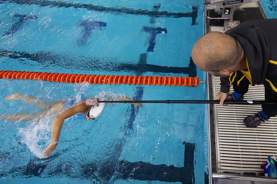 Anastasia Pagonis (kiri) disentuh oleh alat yang dipegang oleh jurulatihnya, Darryn Solotoff semasa salah satu sesi latihan di Pusat Akuatik Nassau County di New York. Acara S11 memerlukan individu khusus untuk menyentuh para atlet sebagai insyarat bila mereka harus berhenti. 