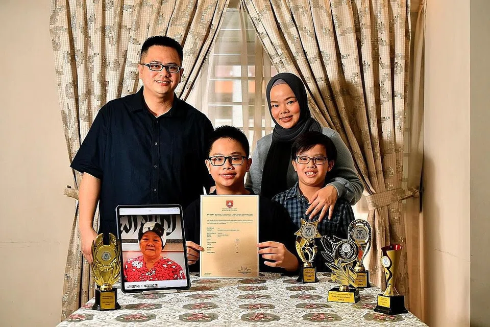 SUKA BERBICARA DALAM BAHASA BERBEZA: Aiman (tengah), yang memegang keputusan PSLE-nya, bergambar dengan ibu bapanya - Encik Faizal dan Cik Nur Azlinda - serta adiknya, Muhammad Aryan Anaqi Mohammad Faizal, lapan tahun. Arwah neneknya, Allahyarhamha Nur Aisyah Brahim (dalam foto berbingkai), banyak mempengaruhi Aiman dan membantunya dalam penguasaan bahasa Melayu sejak kecil. - Foto BH oleh LIM YAOHUI