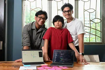 Encik Ilzymanshah Kamarudin (kanan) mereka sebuah alat komunikasi augmentatif dan alternatif (AAC), @Messenger, dengan dibantu adiknya, Encik Irhamshah Kamarudin (kiri), untuk memudahkan komunikasi dengan adiknya, Mohamed Ilhanshah Kamarudin (tengah), yang mengalami gangguan spektrum autisme (ASD).