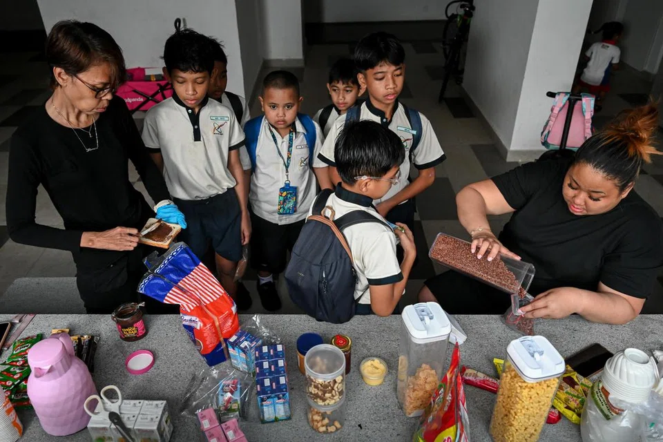 Kanak-kanak bersekolah beratur seawal 6.30 pagi untuk mendapatkan sarapan percuma yang disediakan Cik Lydia Susiyanti Sukarbi (paling kanan) dan rakannya, Cik Halinah Yatim (paling kiri) menerusi inisiatif masyarakat ‘Breakfast Buddy’ yang dijalankan di kolong Blok 228A, Ang Mo Kio Street 23.