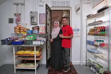 SEDIA MEMBANTU: Cik Norhasyimah Awaludin dan suami Encik Asanul Fariq Sani di  luar pintu rumah mereka yang menyediakan pelbagai barangan runcit untuk kegunaan sesiapa saja yang memerlukan.