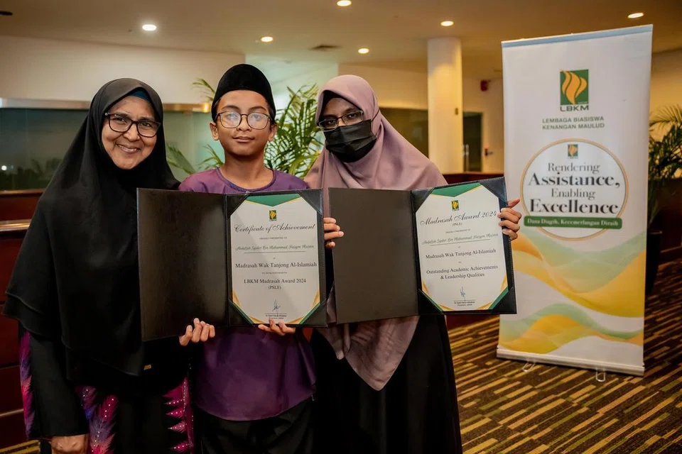 Ibu dan nenek kepada Abdullah Syukri Muhammad Hasyim Masnin, 13 tahun (tengah), iaitu Cik Mardhiyah Md Yusoff (kiri) dan Cik Safiaton Alias (kanan), yang bersinar bangga bersama Anugerah Madrasah LBKM kategori PSLE yang diraih pada 24 Mei lalu.