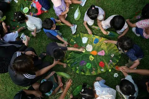 Kanak-kanak dan ibu bapa mereka melukis di atas daun besar dan menghiasinya menggunakan bunga dikumpul mereka semasa meneroka di luar.
