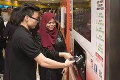 KEDAI CANGGIH: Cik Nur Ainaa Ridzwan, pelajar tahun ketiga Sekolah Pengurusan Perniagaan Politeknik Nanyang, menunjukkan rakan sekolahnya, Encik Nicholas Ong, cara membayar menggunakan telefon bimbit di mesin piza layan diri yang terdapat di kedai serbaneka Cheers tanpa pekerja semalam. - Foto POLITEKNIK NANYANG