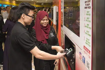 KEDAI CANGGIH: Cik Nur Ainaa Ridzwan, pelajar tahun ketiga Sekolah Pengurusan Perniagaan Politeknik Nanyang, menunjukkan rakan sekolahnya, Encik Nicholas Ong, cara membayar menggunakan telefon bimbit di mesin piza layan diri yang terdapat di kedai serbaneka Cheers tanpa pekerja semalam. - Foto POLITEKNIK NANYANG