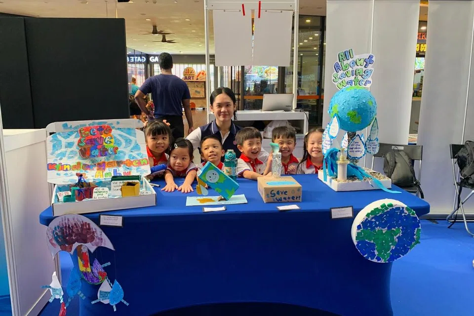 Guru prasekolah di PCF Sparkletots@Jurong Central, Cik Nurul Asyiqin Rahim (tengah), bersama kanak-kanak dan hasil seni mereka di reruai Hari Air Sedunia Singapura 2025. 