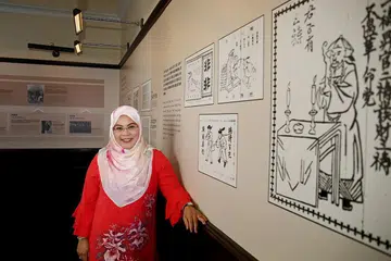 SEMARAKKAN MINAT: Cik Sadiah Shahal menjadi pembimbing muzium bahasa Inggeris bagi SYSMH sejak empat tahun lalu. Beliau menggunakan sejenis kaca mata untuk menjelaskan tentang penglibatan Singapura dalam revolusi China. Komik di kiri Cik Sadiah adalah dari zaman akhir Qing, di mana ia merupakan rakan-rakan revolusi Encik Sun. Ia juga digunakan sebagai bahan publisiti untuk menyebarkan idea-idea mereka dan itulah yang paling beliau minati. - Foto CMG