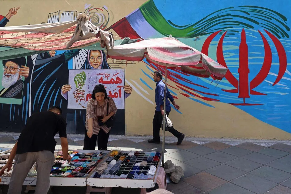 Seorang penjaja jalanan berniaga di hadapan mural di Tehran pada 21 Julai 2026 ketika ketegangan antara Iran dan Amerika Syarikat terus memuncak. Pada hari yang sama, Kor Pengawal Revolusi Islam (IRGC) mendakwa melancarkan serangan ke atas sasaran tentera Amerika Syarikat, termasuk sistem pertahanan udara di Bahrain dan Kuwait, manakala usaha negara pengantara untuk mencapai gencatan senjata selama 11 hari masih diteruskan.