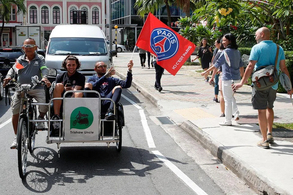 SERONOK JALAN-JALAN: Dua pemain PSG, Adrien Rabiot (tengah) dan Lyvin Kurzawa (memegang bendera PSG) mengambil peluang ketika waktu lapang mereka untuk bersiar-siar mengelilingi Singapura dengan menaiki beca. Mereka juga sempat mencuba beberapa hidangan tempatan seperti membuat prata dan memakan durian. - Foto PSG 