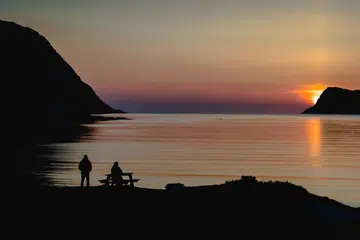 Siluet dua individu berlatarbelakangkan matahari terbenam yang memancarkan cahaya keemasan di atas permukaan air yang tenang.