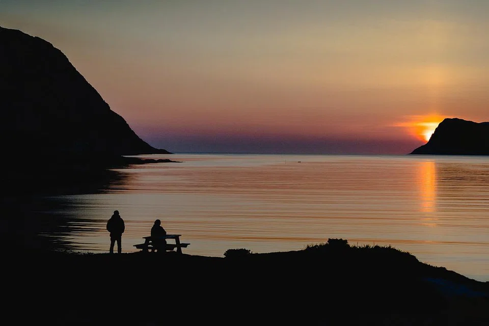 Siluet dua individu berlatarbelakangkan matahari terbenam yang memancarkan cahaya keemasan di atas permukaan air yang tenang.