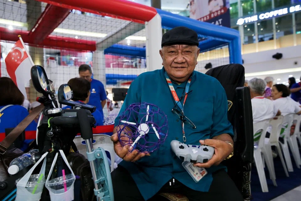 Encik Samsuden Ali, 75 tahun, memegang dron dan alat kawalan semasa menghadiri Kejohanan Bola Sepak Dron Perak Singapura yang pertama di Compass One pada 1 Ogos 2026.
