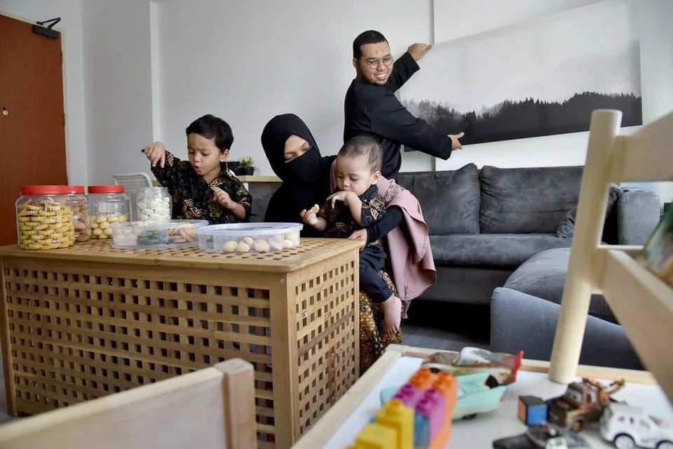 RAYA LEBIH MERIAH: Encik Nor Fahmi Nordin sedang menggantung lukisan bagi menyerikan flat barunya. Isteri, Cik Siti Nur Arfina Peer Mohamed, pula bersama anak-anak mereka, Ahmad Adam (berdiri) dan 'Amr, sedang menjamu kuih-muih, yang sebelum ini tidak dihidangkan di flat sewa mereka. - Foto BH oleh JASMINE CHOONG