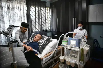 KASIH AYAH: Encik Sunawan (kiri), bapa Encik Asri atau turut dikenali sebagai Riz Sunawan (berbaring), akur berasa sebak melihat anaknya terlantar di katil, namun beliau cekalkan hati supaya dapat membantu anaknya itu menjalani kehidupan. Bersama mereka ialah pembantu rumah yang banyak membantu menguruskan Riz. - Foto BH oleh ARIFFIN JAMAR