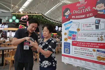 MUDAHKAN PELANGGAN: Seorang rakan digital di cawangan Kopitiam di Jurong Point, Encik Keith Lee, membantu warga emas seperti Cik Peh Ah Eng beralih daripada penggunaan kad fizikal Kopitiam kepada aplikasi FairPrice. - Foto ST