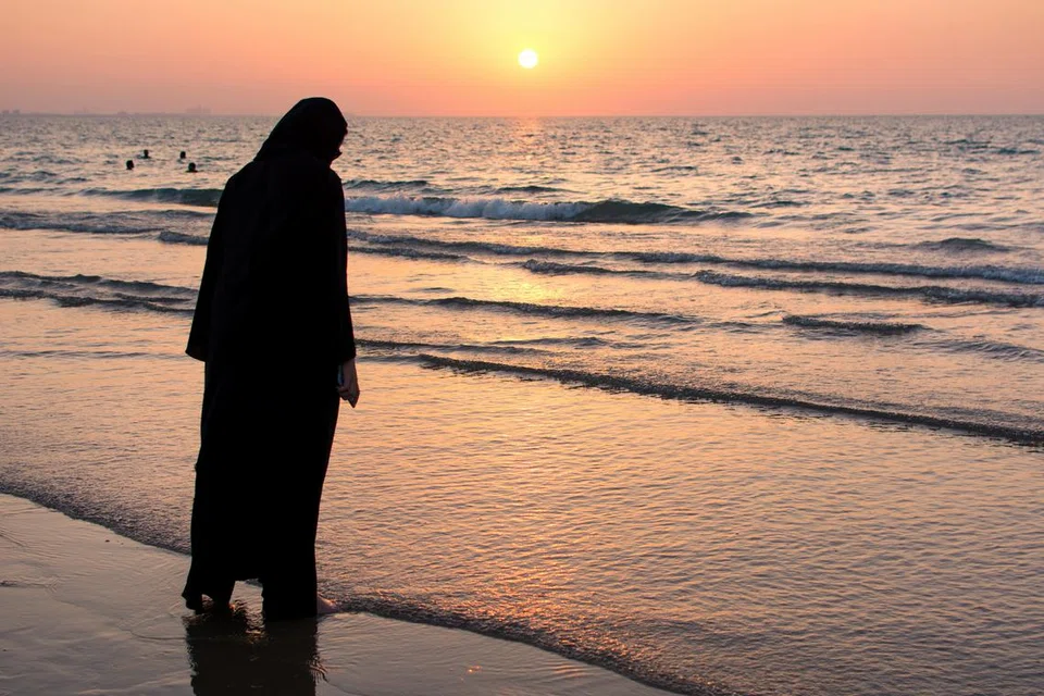 Seorang wanita merenung ke arah matahari terbenam di tepi pantai, menikmati ketenangan dan keindahan alam.