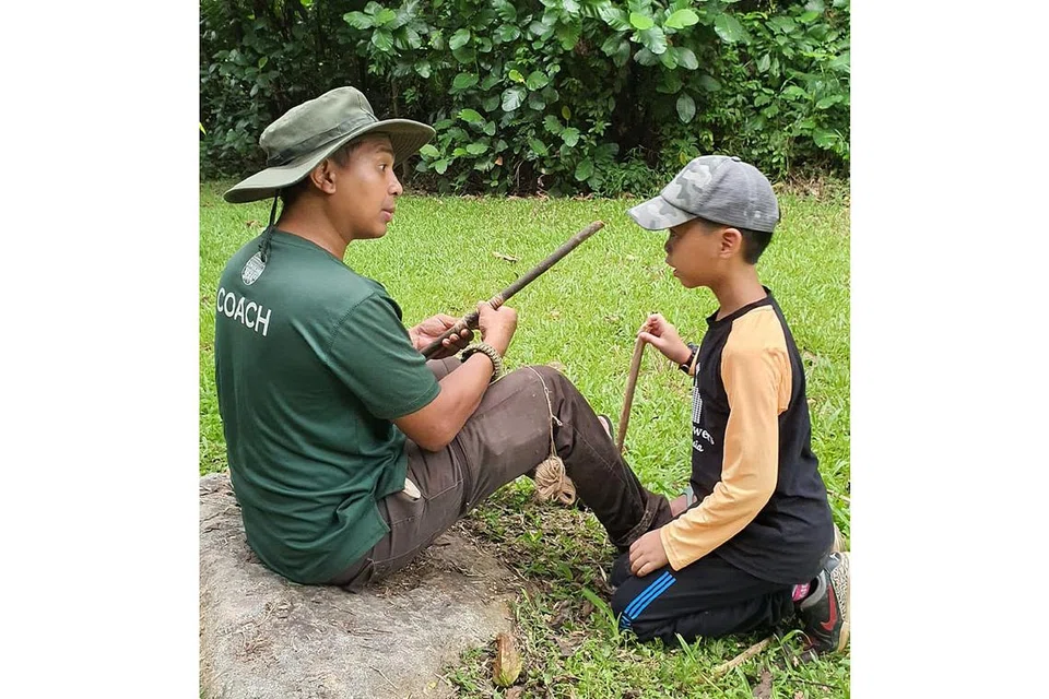 BUDAK KAMPUNG: Pengasas Longkang Adventures, Encik Nor Akasha Abdul Rahman (kiri), bersama salah seorang peserta bagi kem Budak Kampung, selama dua hari satu malam. Encik Nor Akasha sedang mengajar seorang peserta menghasilkan mainan peluru dengan barangan yang didapatkan di hutan. - Foto LONGKANG ADVENTURES