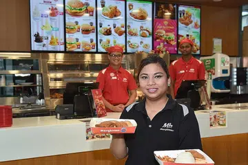 MAKANAN SEGERA: Cik Siti, sedang memegang 'Nasi Lemak MB TM' di tangan kirinya, sementara 'Lucky Plate TM' pula di tangan kanan di hadapan kaunter yang sedang diurus dua kakitangannya, Encik Muhammad Fahmi (kiri), 18 tahun, dan Encik Thinesh Sandrasegaran (kanan), 21 tahun.