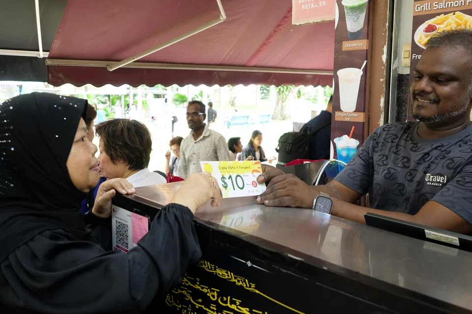Seorang penerima manfaat, Cik Faridah Salamat (kiri), 65 tahun, menebus baucar makan bernilai $10 di kedai makan Mohamed Waseef. Bersamanya ialah pemilik kedai makan Mohamed Waseef, Encik Sathik Ali (kanan).