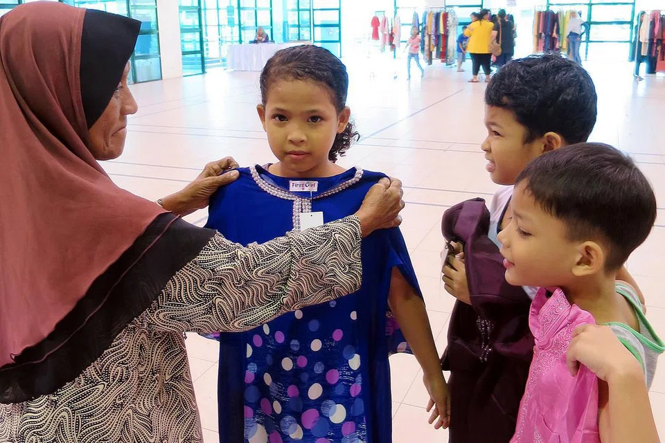 SYUKUR DAPAT BERAYA: Cik Minah Amat (kiri) mengacukan baju kurung biru ke badan cucunya, Nur Atrisya Zainudin, semasa pengagihan baju Hari Raya anjuran Angkatan Karyawan Islam (AMP) dan Second Chance di Kelab Masyarakat Pasir Ris East pada Sabtu. Turut bersama mereka dua lagi cucunya, Muhamad Hafiz Zainudin (dua dari kanan) dan Muhamad Fukaliman Zainudin. - Foto AMP
