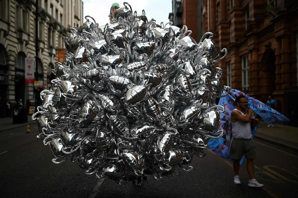 REBUT PELUANG: Seorang penjual belon berbentuk trofi tiba awal di kawasan yang bakal dilalui perarakan pemain Manchester City bagi mendapatkan tempat terbaik untuk menjual belon-belonnya. Usahawan kecil seperti lelaki ini membanjiri sekeliling jalan raya Manchester semasa perarakan bas di Manchester City pada 12 Jun. 