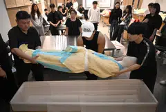 Busan Institute of Science and Technology students lowering a mannequin dressed in traditional Korean funeral cloth into a coffin during a funeral administration class.