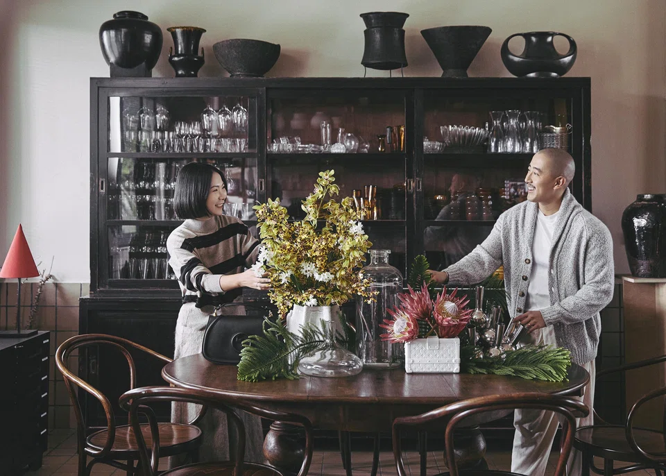 Posing at This Humid House, Lilian Sun wears a striped cashmere pullover and mohair skirt by Brunello Cucinelli. Jonathan Wong wears white cotton T-shirt, cashmere knitted cardigans and off-white corduroy trousers by Brunello Cucinelli. Tableware includes Petite Malle Porcelain Vase and Porcelain Vase Noe BB by Louis Vuitton. Floral design by This Humid House.