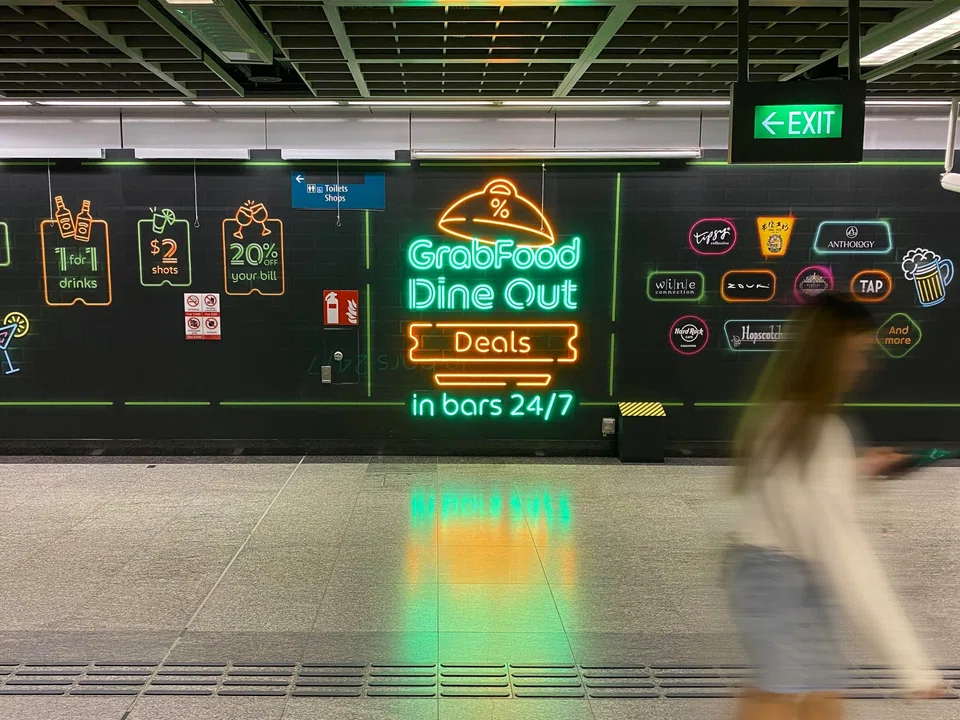 Grab's buzzy, neon-lit sign announcing “GrabFood Dine Out Deals in bars 24/7” at Clarke Quay MRT station.