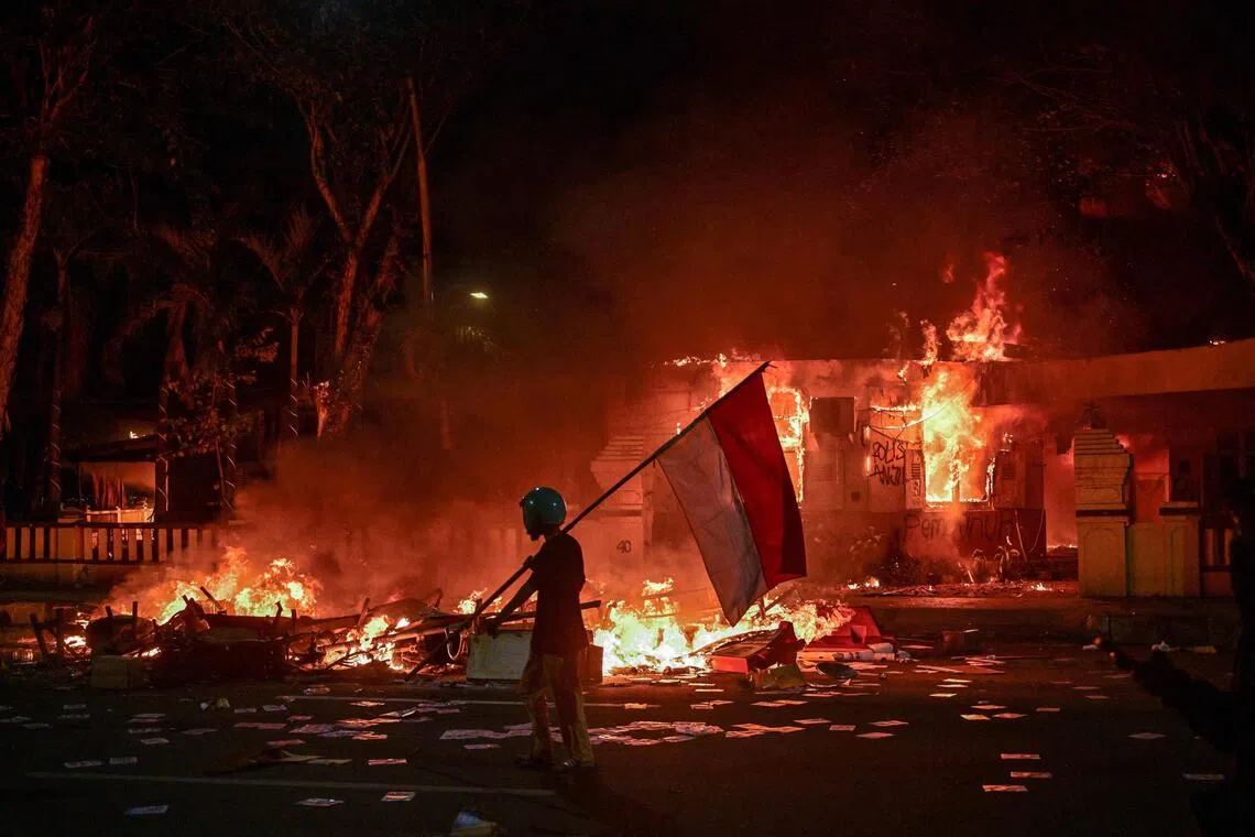 A protester walks with an Indonesian flag in front of a police headquarters that was set on fire and looted during demonstrations in Surabaya on August 31, 2025. Southeast Asia's biggest economy has been rocked by protests in major cities, including the capital Jakarta, since April 29 after footage spread of a motorcycle taxi driver being run over by a police tactical vehicle during an earlier rally against low wages and financial perks for lawmakers. (Photo by Juni KRISWANTO / AFP)
