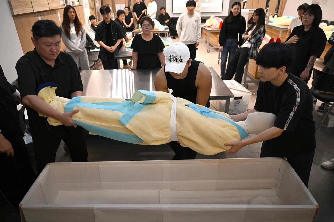Busan Institute of Science and Technology students lowering a mannequin dressed in traditional Korean funeral cloth into a coffin during a funeral administration class.