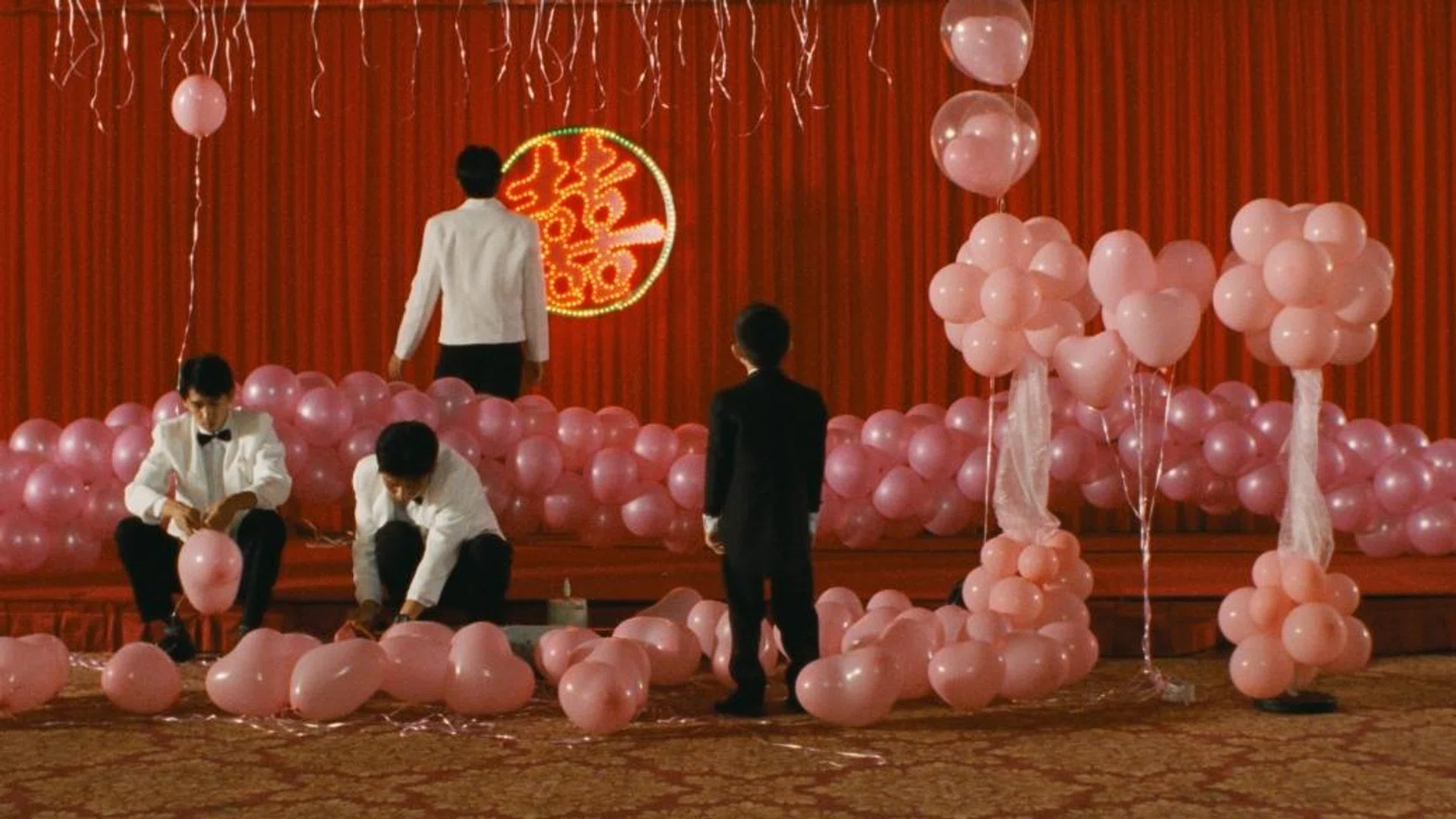 Still from Edward Yang’s film Yi Yi showing children and adults arranging pink balloons on a stage with red curtains, capturing a quiet, ceremonial moment from the Taiwanese family drama.