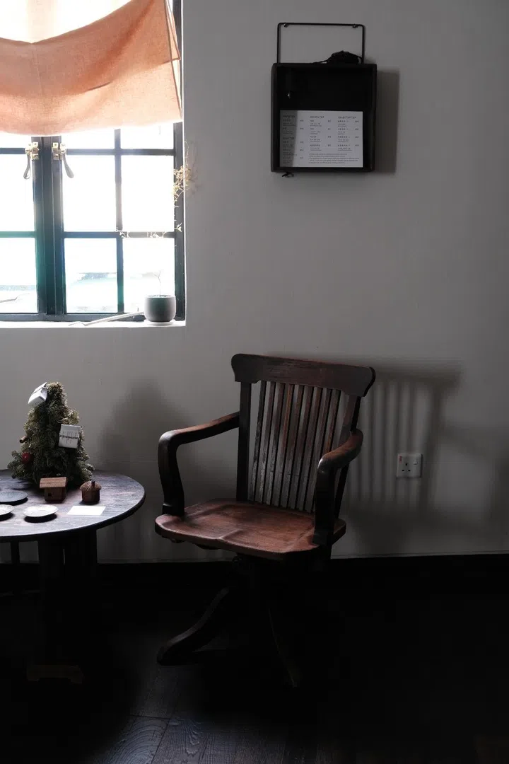 Sunlit window corner at Inner Teahouse in Chinatown Singapore featuring a vintage wooden chair and cafe table under a soft linen blind with a view of the surrounding HDB and shophouse architecture.