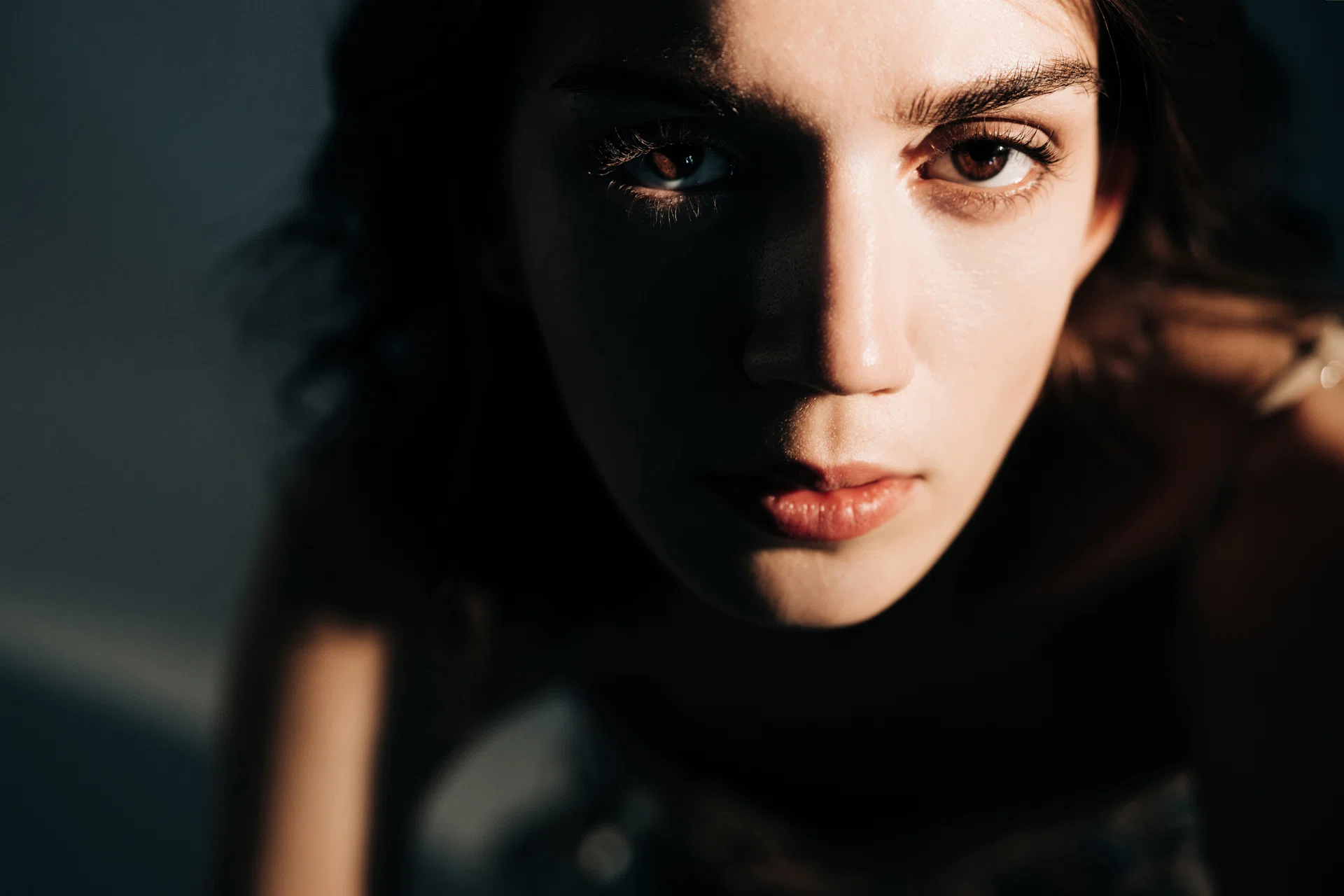 A close-up portrait of a woman in dramatic light, with shadows emphasizing her eyes and facial features. The intimate framing conveys emotion, depth, and cinematic allure.